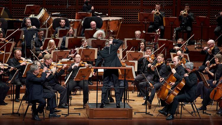 A wide shot of musicians performing under a conductor’s direction on a concert hall stage