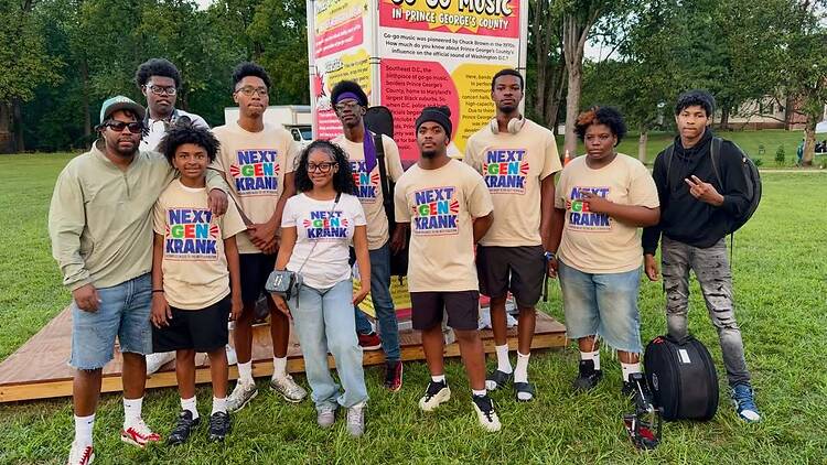 A wide shot of a group posing together outdoors in front of a Go-Go Music exhibit. A wide shot of a group posing together outdoors in front of a Go-Go Music exhibit.
