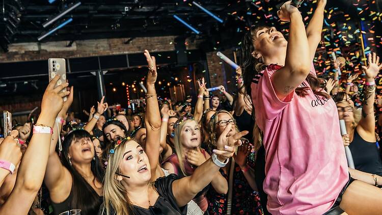 A wide shot of a singer performing into a microphone as a crowd cheers and confetti falls. A wide shot of a singer performing into a microphone as a crowd cheers and confetti falls.