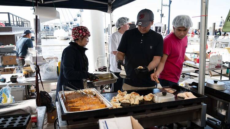 A wide shot of vendors preparing food at an outdoor market by the waterfront. A wide shot of vendors preparing food at an outdoor market by the waterfront.