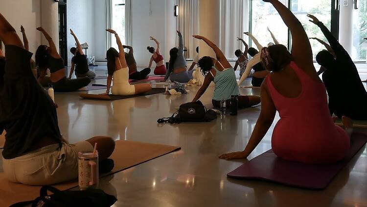 A wide shot of people practicing a seated side stretch together in a yoga studio. A wide shot of people practicing a seated side stretch together in a yoga studio.