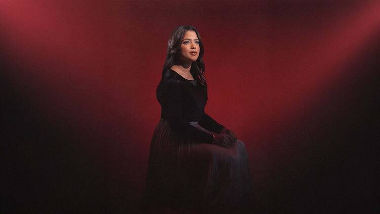 A medium shot of a person seated and posing against a red backdrop. A medium shot of a person seated and posing against a red backdrop.