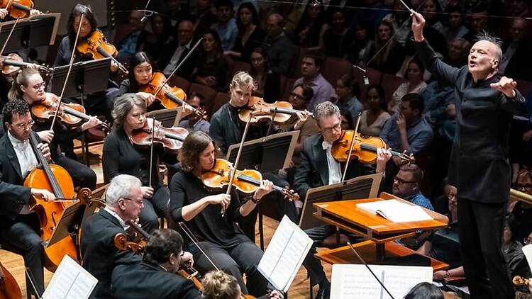 A wide shot of an orchestra performing with a conductor leading the musicians on stage. A wide shot of an orchestra performing with a conductor leading the musicians on stage.