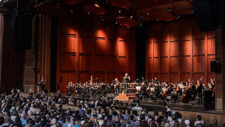 A wide shot of an orchestra performing with a conductor on stage before a seated audience. A wide shot of an orchestra performing with a conductor on stage before a seated audience.
