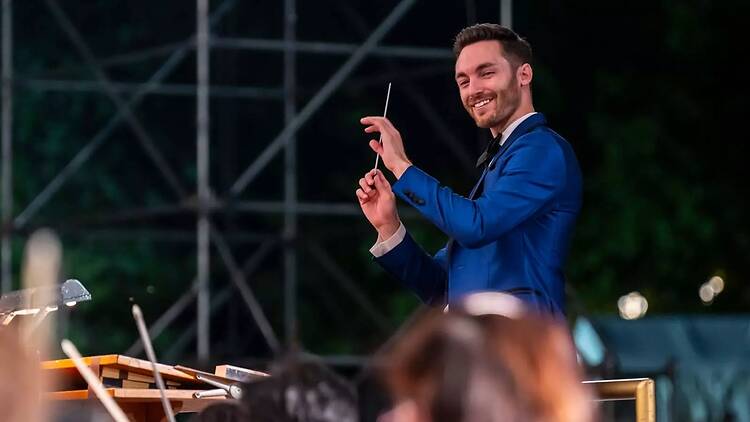 A wide shot of a conductor leading an orchestra during an outdoor performance.