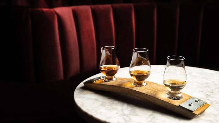 A close-up of three whiskey glasses arranged on a wooden tasting board on a table. A close-up of three whiskey glasses arranged on a wooden tasting board on a table.