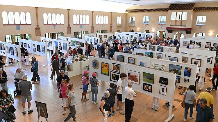 A wide shot of visitors browsing artwork displayed at a large indoor art exhibition. A wide shot of visitors browsing artwork displayed at a large indoor art exhibition.