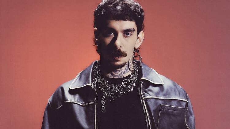 A close-up of a person wearing a leather jacket and layered chains against a red backdrop. A close-up of a person wearing a leather jacket and layered chains against a red backdrop.