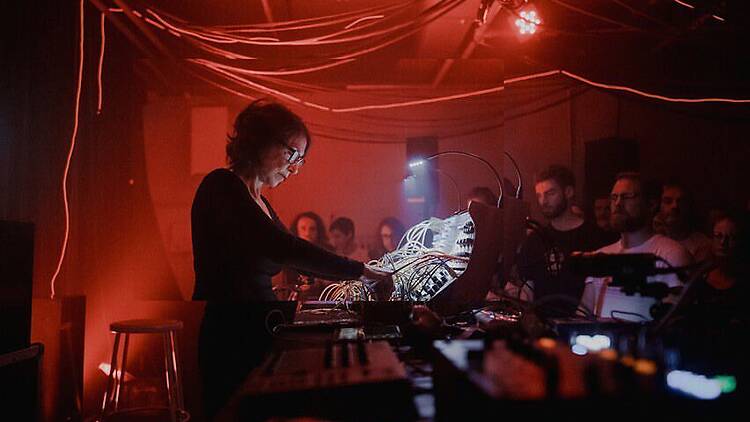 A wide shot of a DJ performing with electronic equipment as people watch in a dimly lit venue. A wide shot of a DJ performing with electronic equipment as people watch in a dimly lit venue.