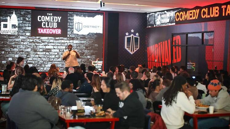 A wide shot of a comedian performing on stage for an audience at a comedy club. A wide shot of a comedian performing on stage for an audience at a comedy club.