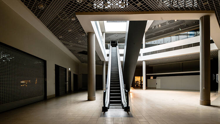 Stinsen, Sollentuna Empty mall corridor in the shuttered Stinsen shopping centre. An escalator leads up to an upper level, shop units sit closed behind metal shutters, and daylight falls from a skylight above.