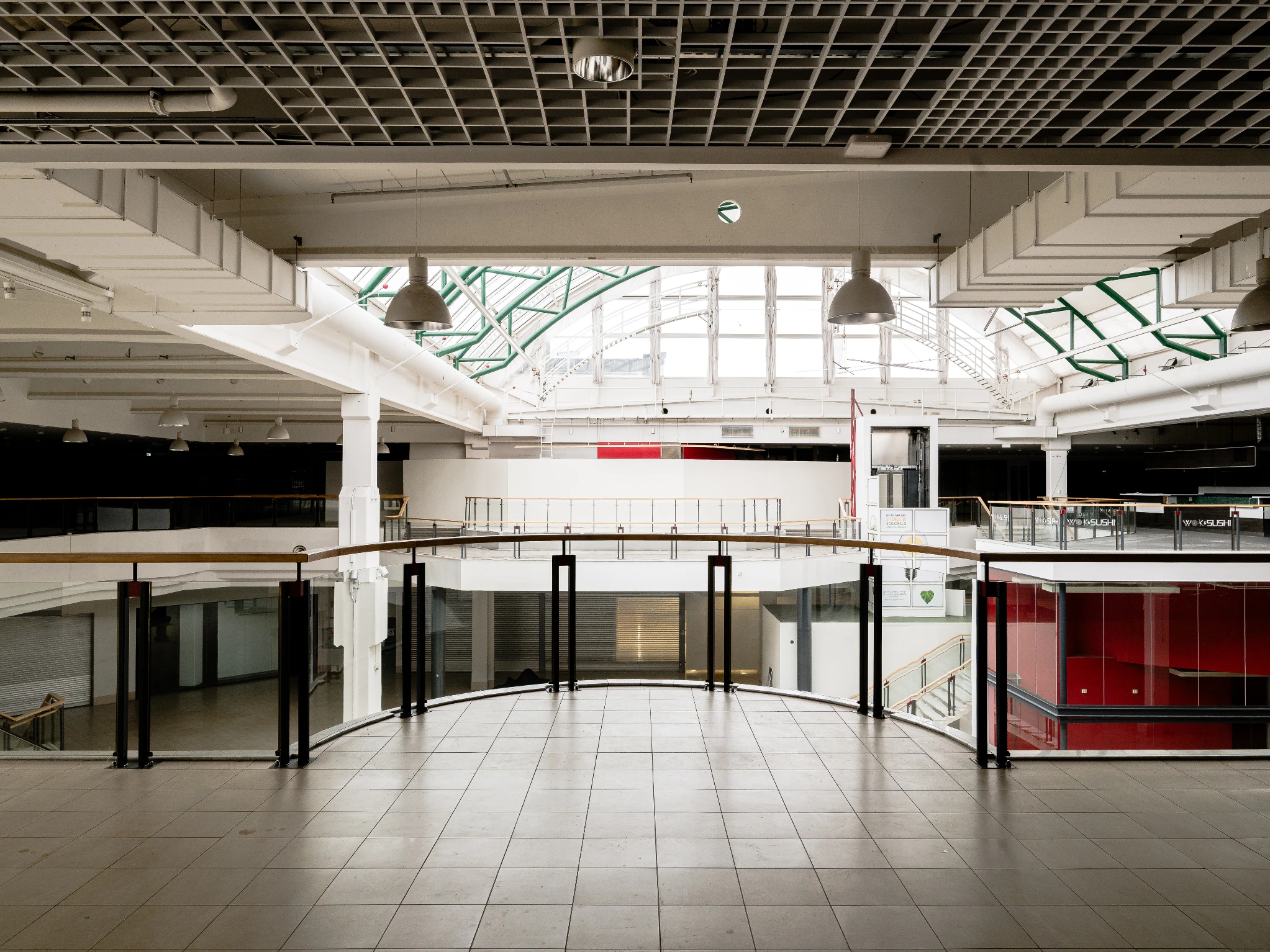 Empty mall corridor in the shuttered Stinsen shopping centre. An escalator leads up to an upper level, shop units sit closed behind metal shutters, and daylight falls from a skylight above.