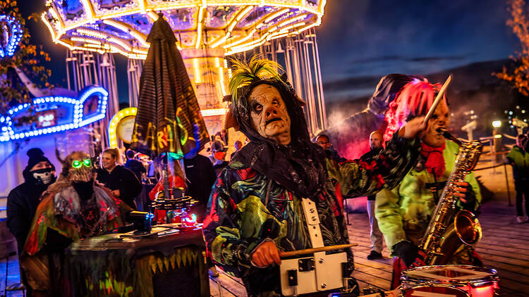 Halloween Gröna Lund Costumed musicians perform outdoors at night during Halloween at Gröna Lund. A drummer in a mask and ragged coat stands in the foreground, with a saxophonist in a green outfit and an illuminated carousel behind.