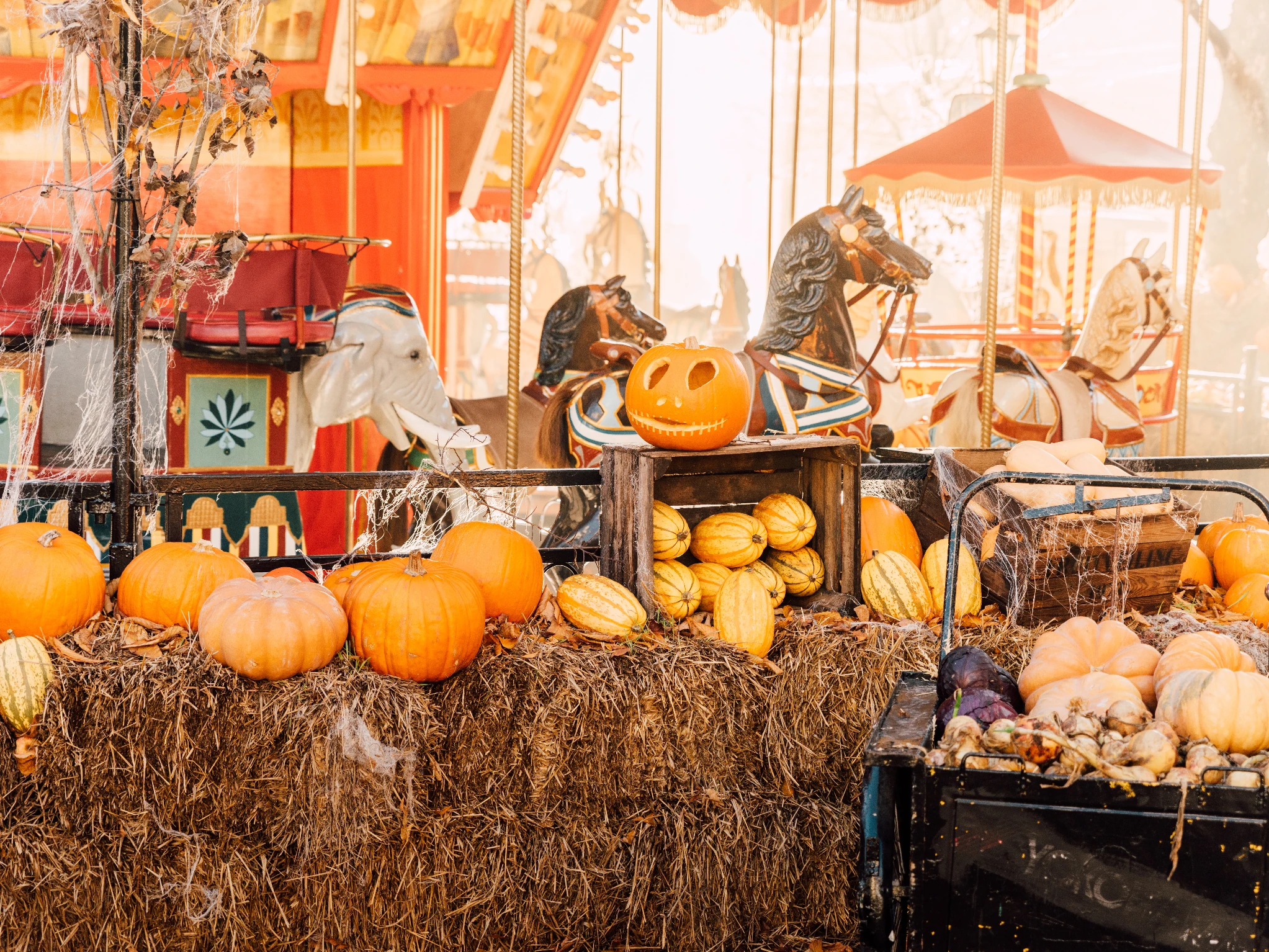 Halloween decorations at Gr&ouml;na Lund: pumpkins, squash and wooden crates arranged on straw bales, with a carved jack-o'-lantern on top and a carousel with painted horses behind.