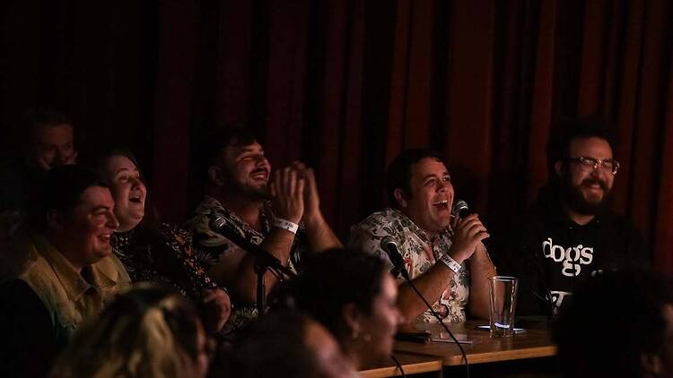 A wide shot of people seated at a table speaking into microphones during a live comedy show