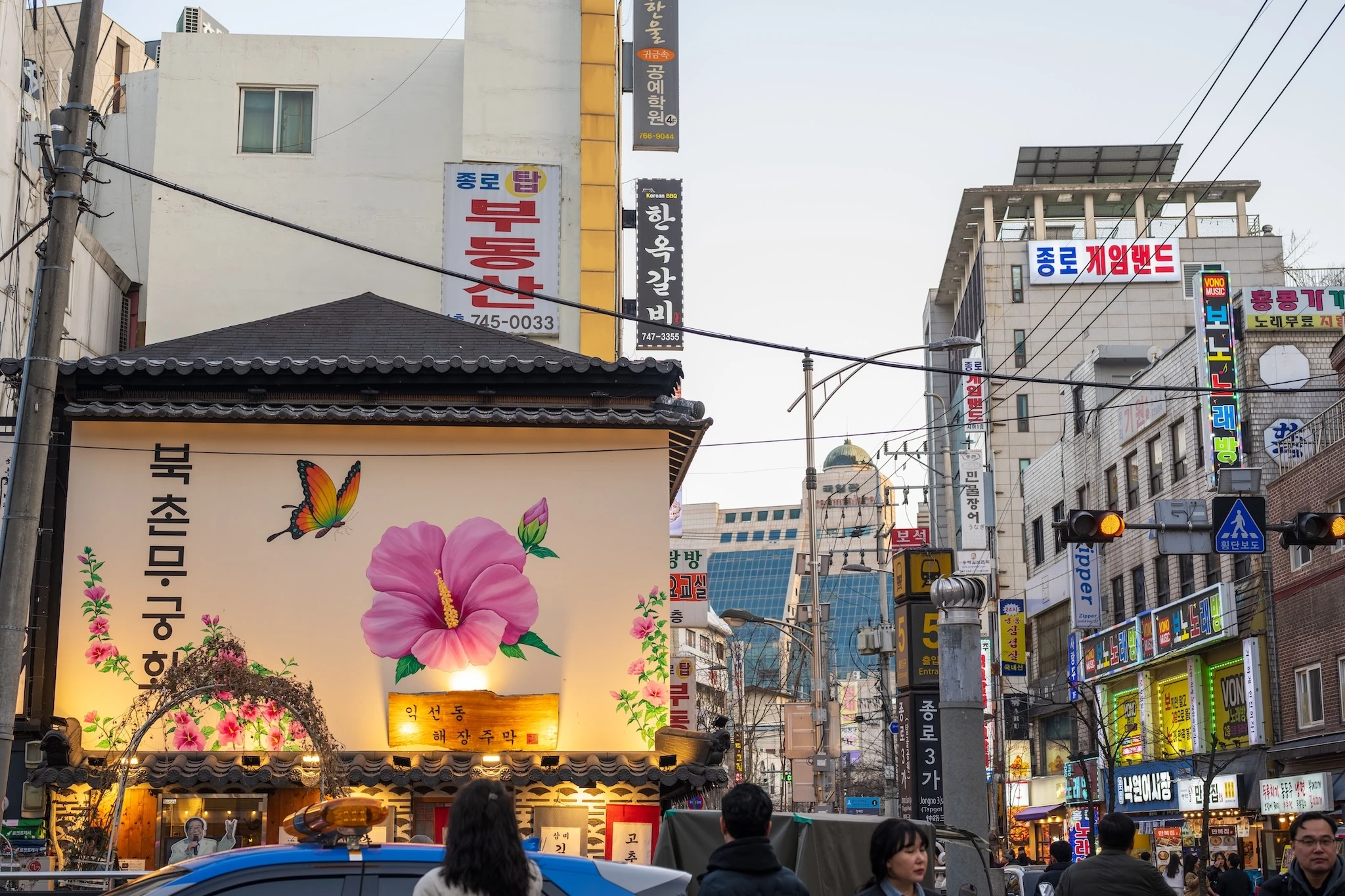 A traditional tiled building with a large flower mural stands amid a dense cluster of neon signs and businesses near Jongno 3-ga station