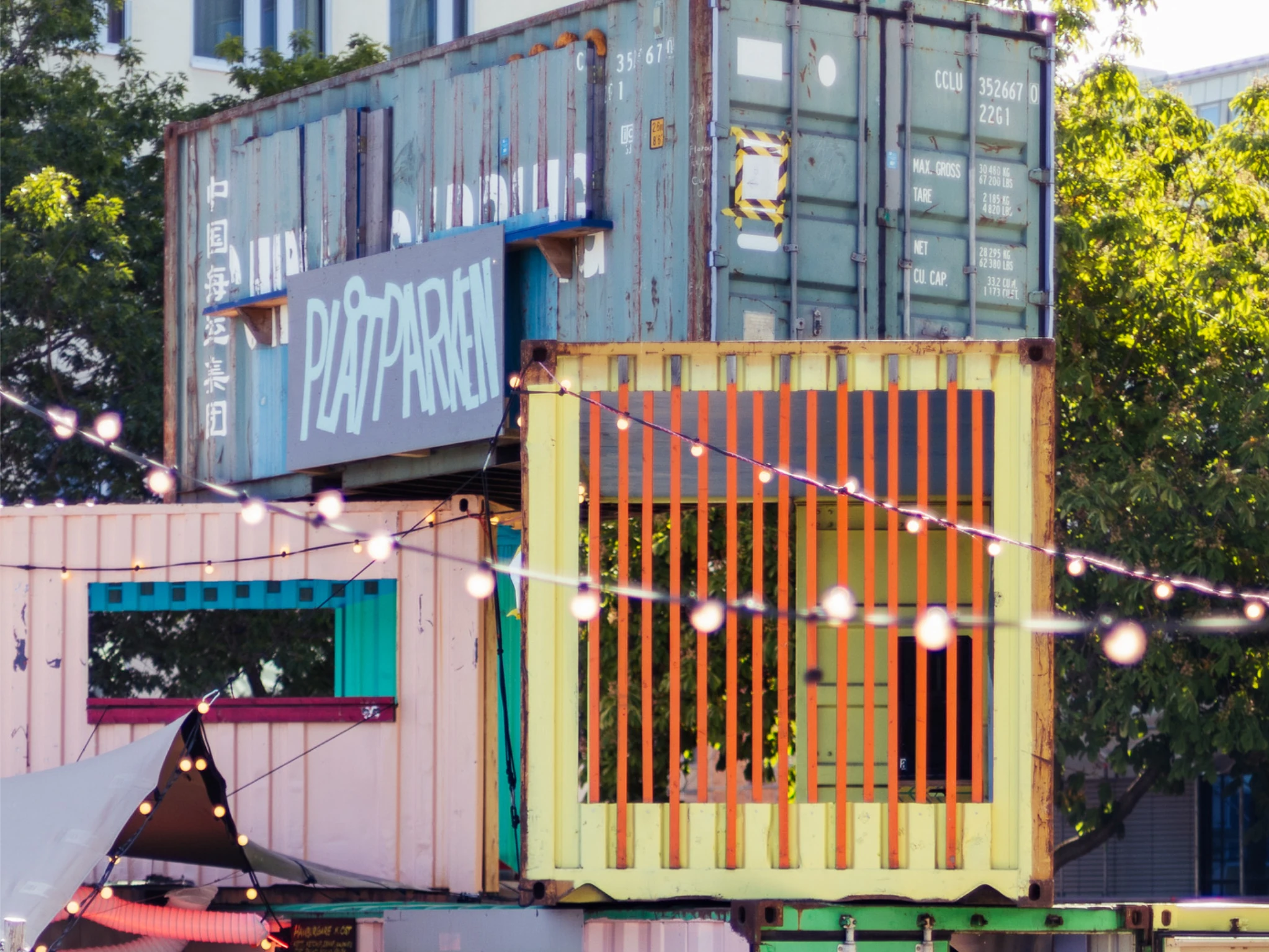 Stacked shipping containers in pastel colours with a hand-painted "Pl&aring;tparken" sign, strung with festoon lights and framed by trees.
