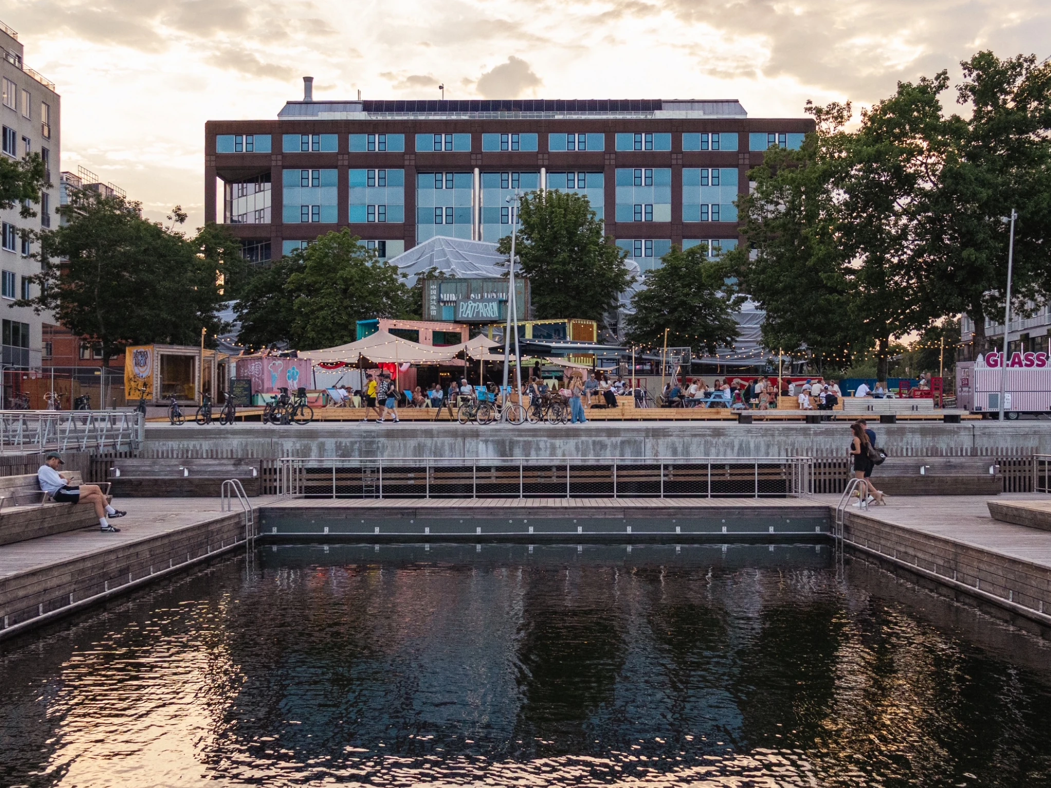   Marievik's quayside swimming pool at dusk, with Pl&aring;tparken's containers, awnings and festoon lights busy with people behind it.
