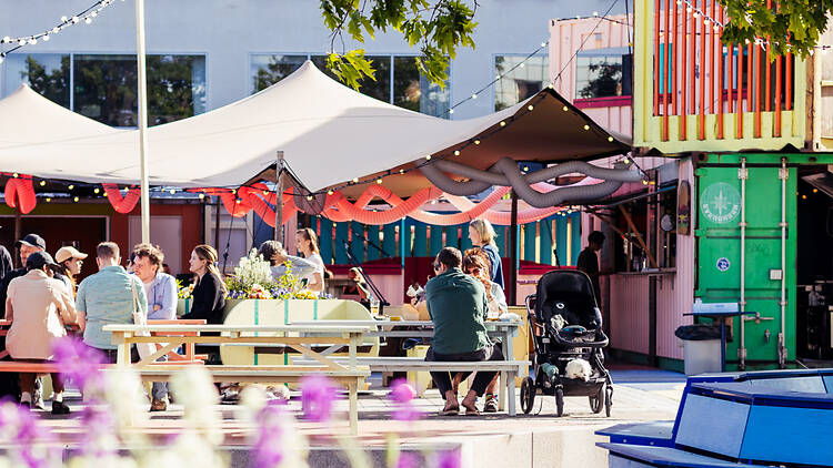 Plåtparken, Marievik, Stockholm Groups eating at picnic tables under canvas awnings and festoon lights at Plåtparken, surrounded by brightly painted shipping containers.