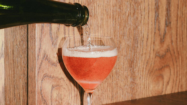 Dopp, Östermalm, Stockholm A dark green bottle pouring sparkling wine into a stemmed glass already filled with a pink drink, topping it with a layer of foam. The glass stands against a pale wooden panel wall.