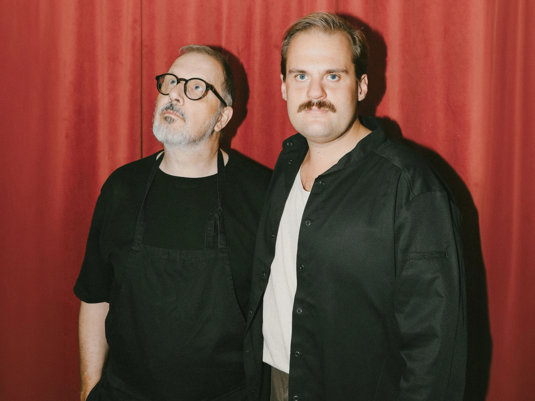 Two men in black standing against a red curtain, one in round glasses and an apron looking upward, the other looking straight at the camera.
