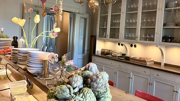 Open kitchen dining area at Ett Hem hotel in Stockholm, featuring fresh produce like artichokes, tulips, glass-front cabinets, and a brass chandelier.