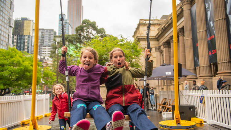 SWING two girls sit on a giant yellow swing with a third girl behind them