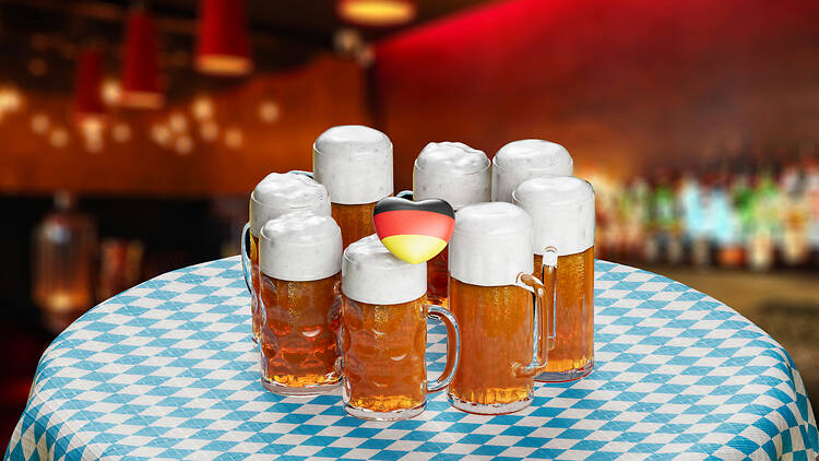 Eight full beer steins with thick foam heads on a blue and white chequered Bavarian tablecloth, with a small German flag heart among them and a blurred bar behind. Eight full beer steins with thick foam heads on a blue and white chequered Bavarian tablecloth, with a small German flag heart among them and a blurred bar behind.