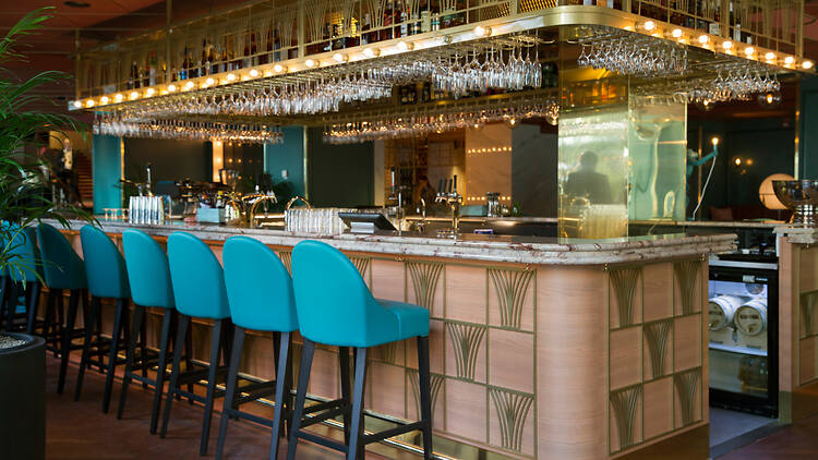 Stockholm, Haymarket Americain Cocktailbar An art deco bar with turquoise stools, a marble counter and a brass overhead rack of hanging wine glasses.