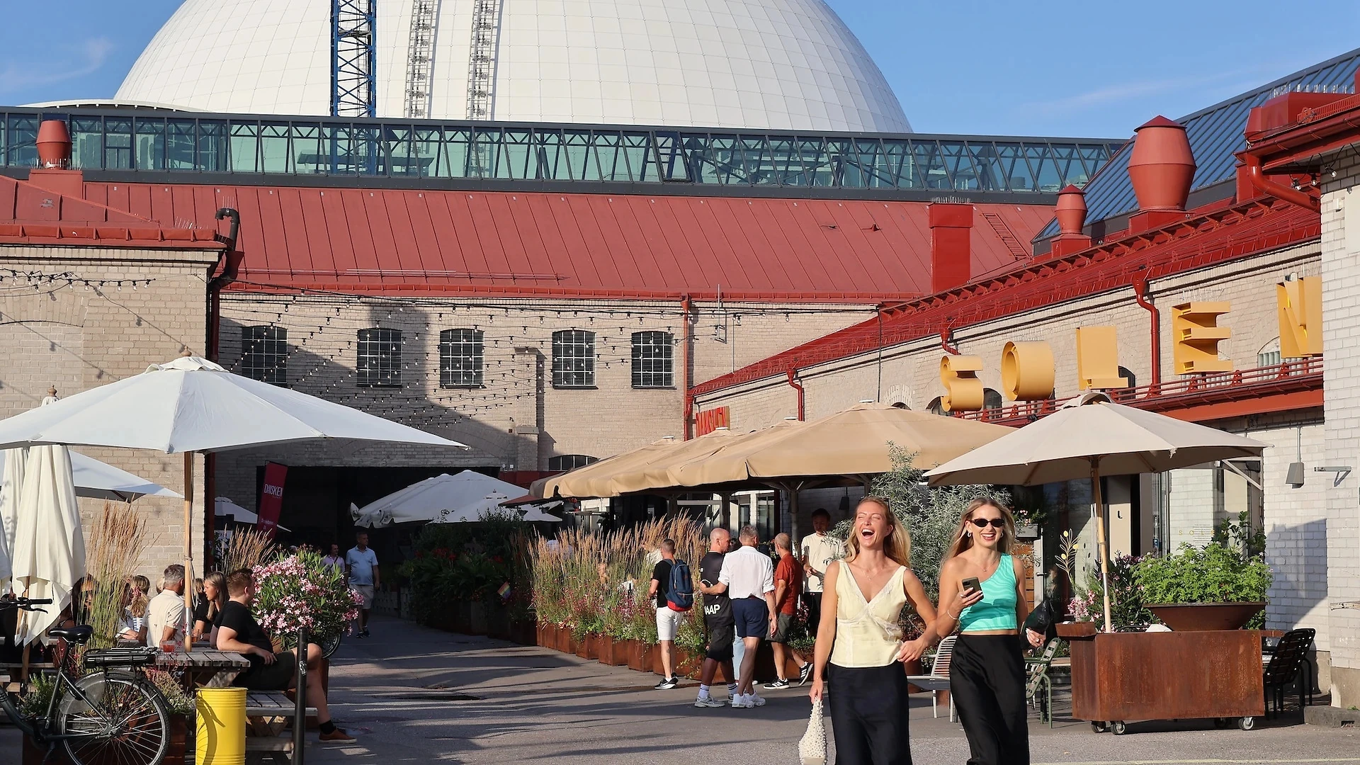 kaksi naista hymyilee etualalla, taustalla teollisuusrakennus ja sen takana Globen-halli