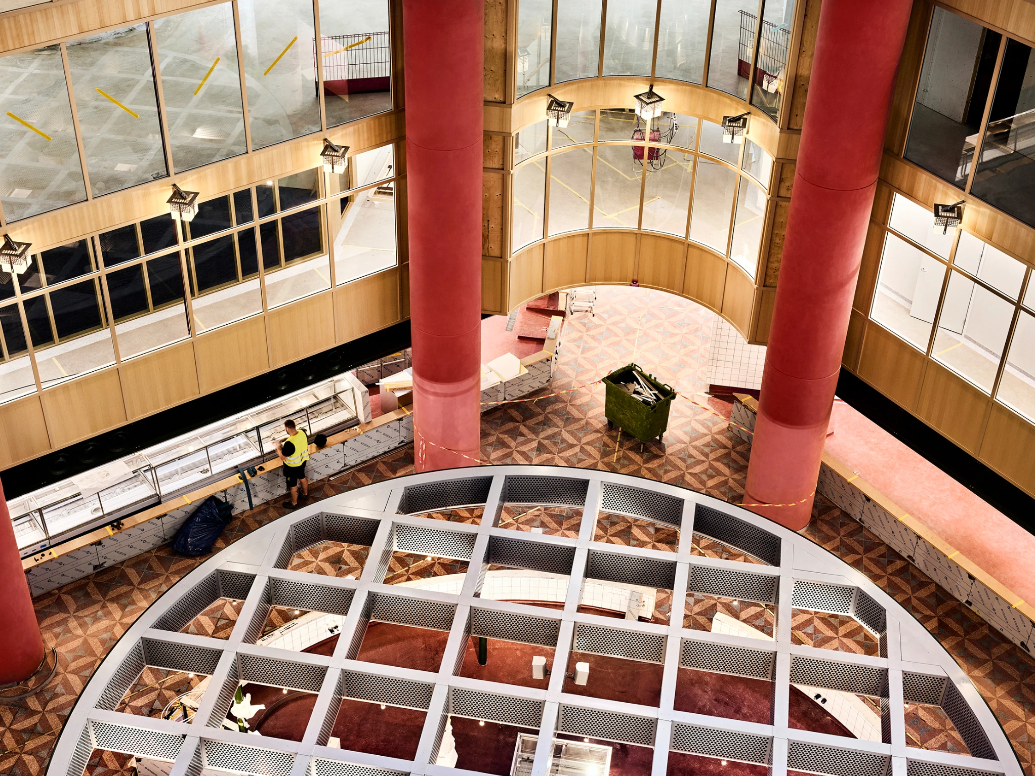 An overhead view of S&ouml;derhallarna under renovation, with red columns, wrapped market counters and a worker on the patterned floor.