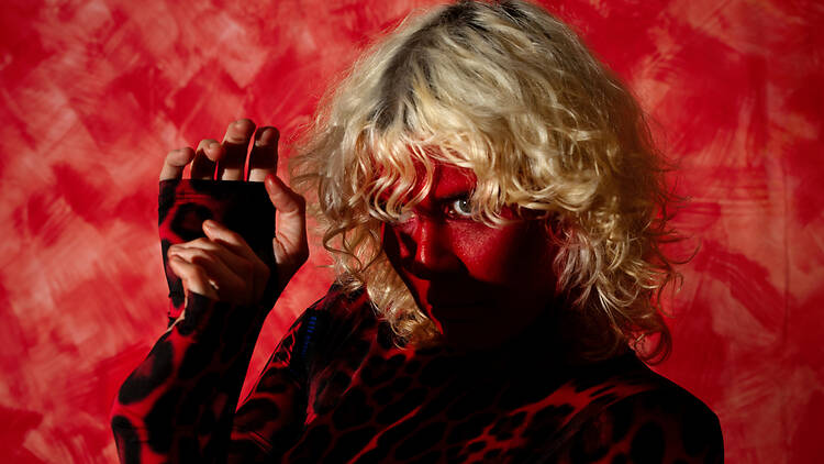 Sibille Attar Pressbild Sibille Attar in red face makeup and a red leopard-print top, posing against a red painted backdrop.