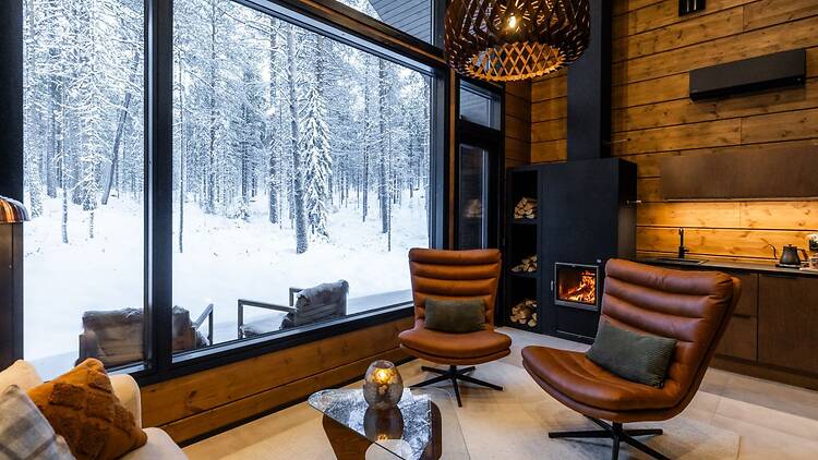 Cozy modern cabin interior with floor-to-ceiling windows overlooking a snowy forest, featuring leather lounge chairs, a fireplace, and warm wooden finishes. Cozy modern cabin interior with floor-to-ceiling windows overlooking a snowy forest, featuring leather lounge chairs, a fireplace, and warm wooden finishes.