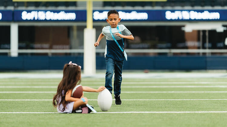 A young boy running to kick a football held by a little girl on the field in SoFi Stadium