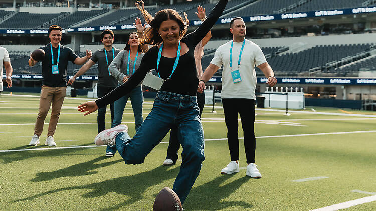 A woman kicking a football off a tee in front of a small group on the field of SoFi Stadium