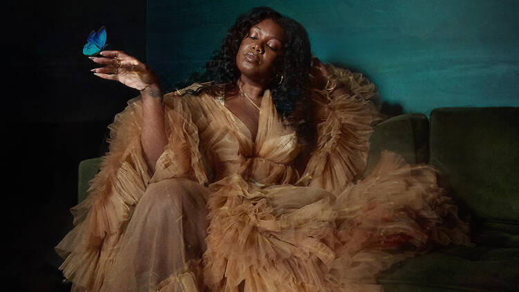 Yasmin Lacey, jazzartist Yazmin Lacey, eyes closed, sits on a green velvet sofa in a flowing ruffled tulle gown, a blue butterfly resting on her raised hand, beneath a candle-style wall sconce in a dark teal room.