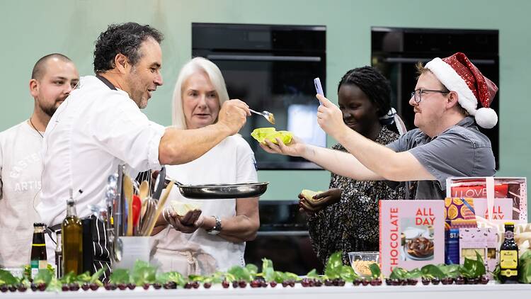 Good Food & Wine Show Christmas Edition a chef with a frying pan serves a spoon of food to a man wearing a Santa hat