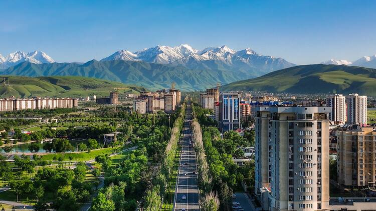 Aerial panoramic view of a wide, tree-lined street and modern buildings in Bishkek with the snowy Tian Shan mountains in the distance Aerial panoramic view of a wide, tree-lined street and modern buildings in Bishkek with the snowy Tian Shan mountains in the distance