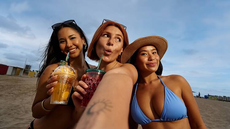 Three women on holiday in Barcelona Three young adult women enjoying beach vacation, smiling and holding drinks while taking a selfie with sand and sky