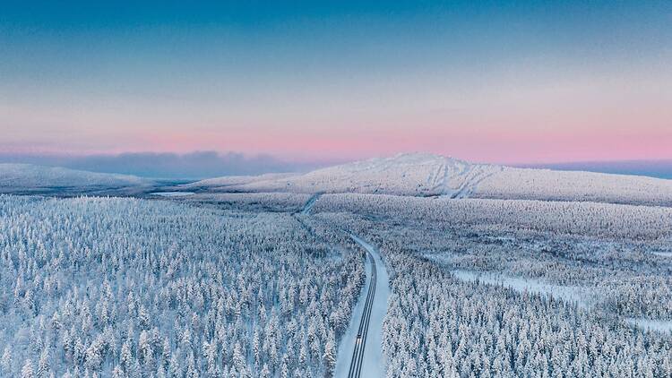 Snow-covered forests and rolling fells in Finnish Lapland, with a winding winter road stretching toward the horizon beneath a pastel Arctic sky.