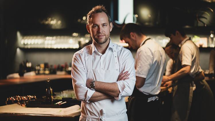 Chef Björn Frantzén stands with his arms crossed in a white shirt and white apron, looking into the camera. Behind him, in a softly lit restaurant kitchen, three cooks are busy at a counter in front of shelves lined with glasses and bottles.