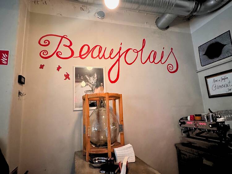 Interior Wall Art at Ninja Wine Bar in Stockholm A wall featuring red script lettering that reads Beaujolais above a framed cat picture and a glass demijohn in a wooden crate.