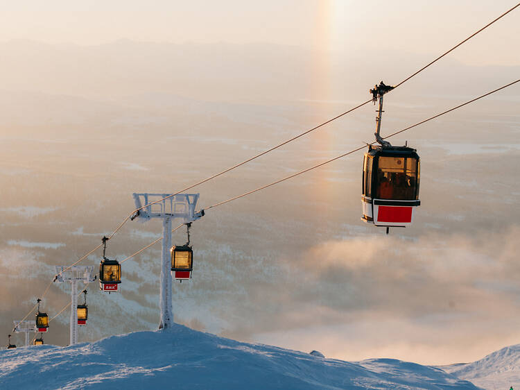 : best things to do in Åre: Ride the Cable Car and Gondola up Åreskutan Mountain Cable car gondolas carrying skiers up snowy Åreskutan mountain against a hazy winter sunset background in Åre, Sweden.