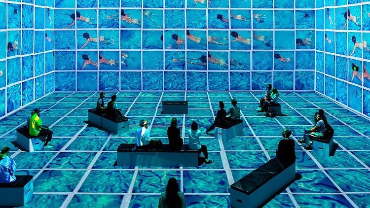 David Hockney: Bigger & Closer (not smaller & further away) people sitting down surrounded by blue screens to represent a swimming pool with people swimming