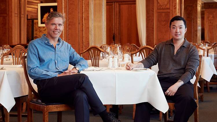 Two men sit at a white-clothed table in Bobergs’ wood-panelled dining room at NK. Around them are wine glasses and rows of set tables.