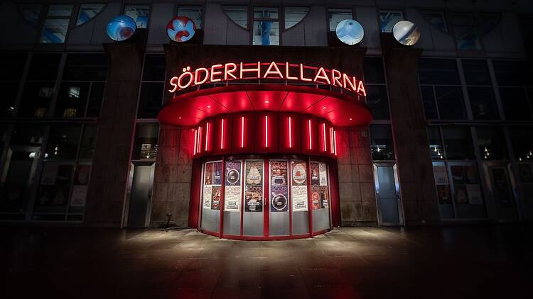 The entrance to Söderhallarna at night: a red neon sign above a curved, red-lit doorway covered in posters announcing the reopening on 23 October.