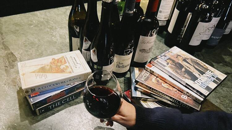 hand holding a glass of red wine over a stone bar counter, next to stacks of VHS tapes and DVDs and a row of wine bottles.