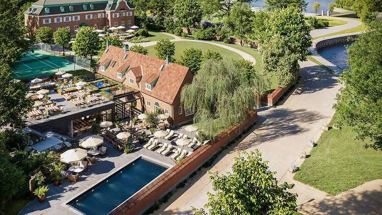 Aerial view of Villa Pauli in Djursholm, with the brick villa, a tennis court, rooftop terraces, a pool lined with sun loungers and parasols, and the water in the background.